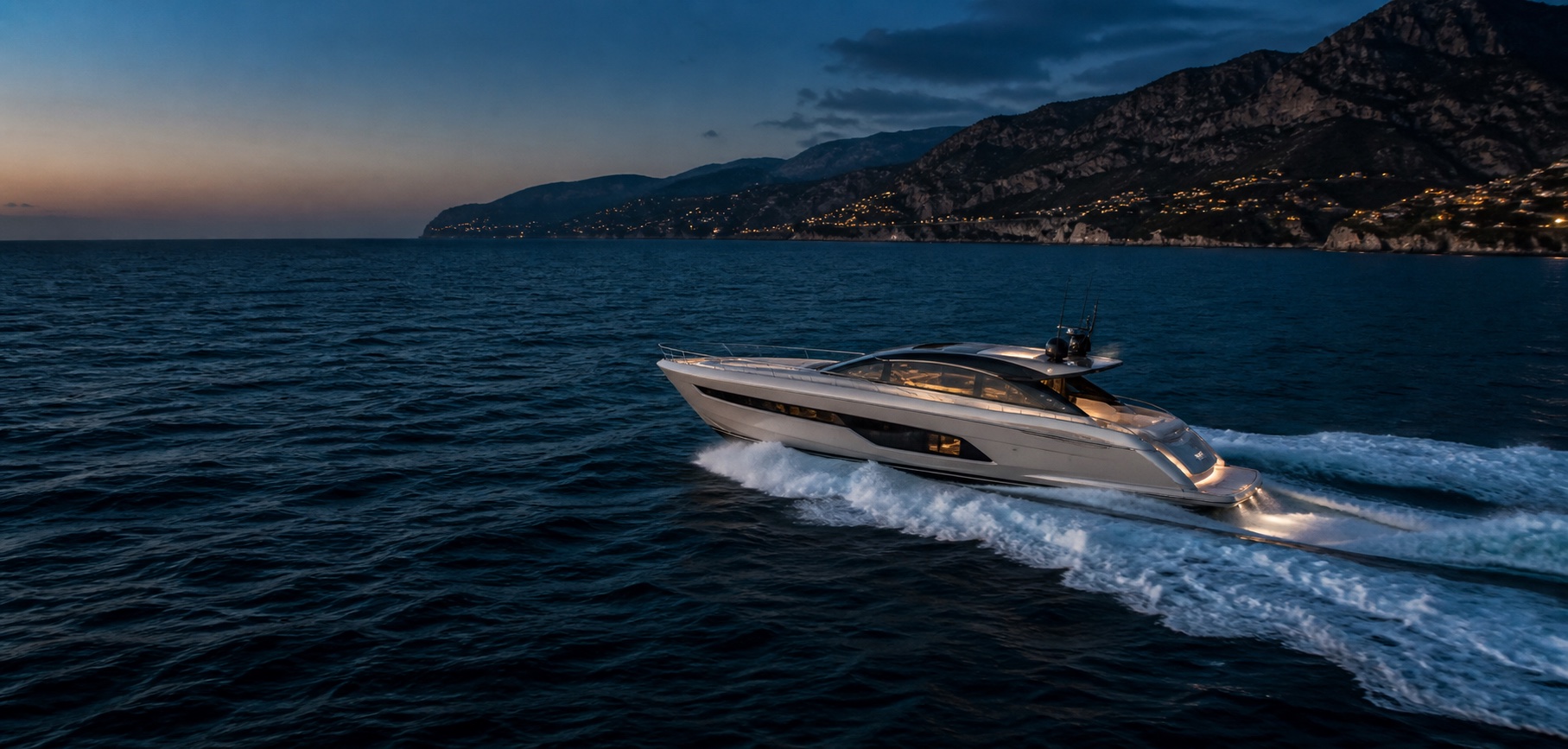 A performance motor yacht travelling along a Mediterranean coastline at blue hour