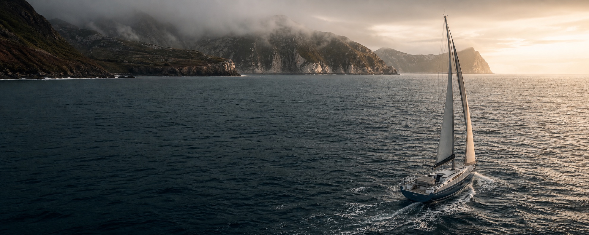 A blue-water sailing yacht making steady progress past a misty European coastline at dawn