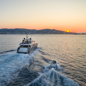A large yacht with a smaller tender boat trailing behind it, sailing into the sunset with hills on the horizon