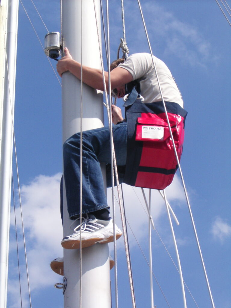 Person aloft beside a sailboat mast in a bosun’s chair